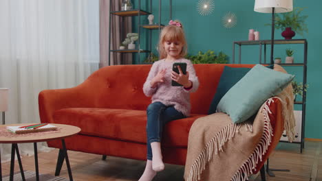 Little-toddler-school-kid-girl-taking-selfie-photo-with-smartphone-while-sitting-on-sofa-at-home