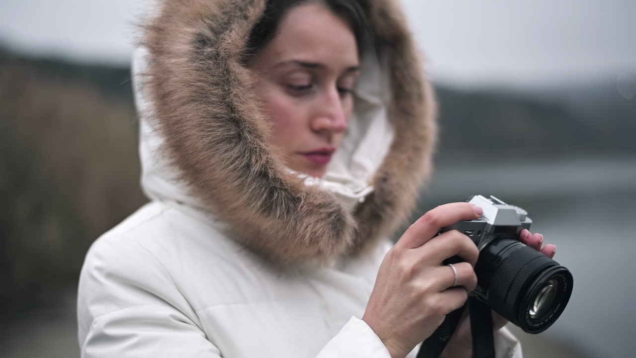 A woman wearing a warm white coat with a fur hood carefully adjusts her camera while standing by calm water. The cloudy sky adds a soft ambiance to the chilly winter scenery