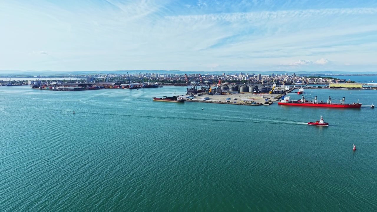 Scena marittima ripresa. Porto industriale con navi e gru attive oggi. Vista aerea che mostra rimorchiatori guidare una petroliera in un vivace ambiente portuale, ricco di macchinari.