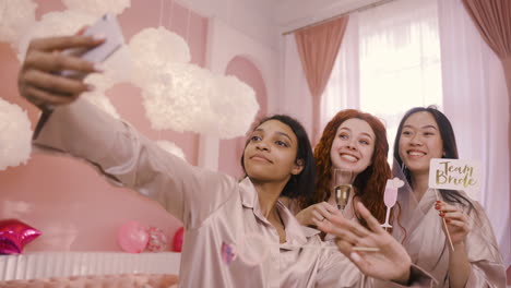 Three-Multiethnic-Friends-Holding-Cardboard-Decoration-And-Crystal-Glasses-Of-Champagne-Taking-A-Selfie-With-Smartphone-In-Bachelorette-Party-2