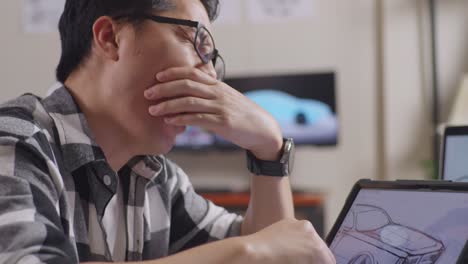 close up of asian male yawning and sleeping while drawing new car design concept on a tablet in the studio with tv and computers display 3d electric car model