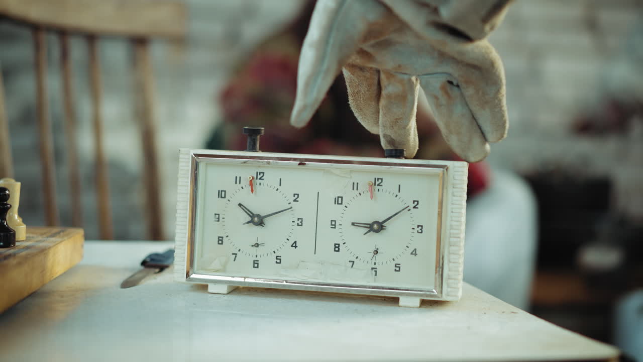 Close up of hand in glove adjusting chess clock on table with blurred people in background inside shelter, symbolizing passage of time, and resilience during crisis in difficult living conditions