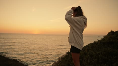 young girl in sweatshirt touching hair on sea cliffs at sunset