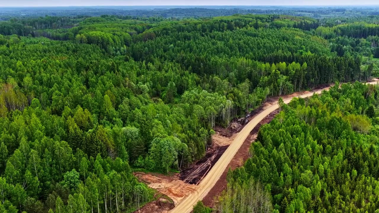 Filmisk luftoptagelse af en ny grusvej, der bliver anlagt gennem en frodig grøn skov til et bæredygtigt vindmølleprojekt i Letland.