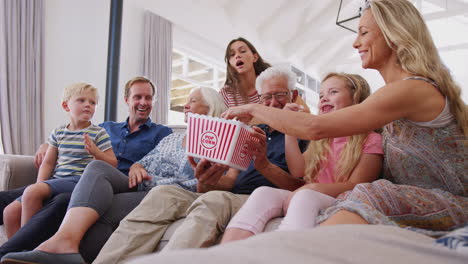 multi-generation family sitting on sofa at home eating popcorn and watching movie together