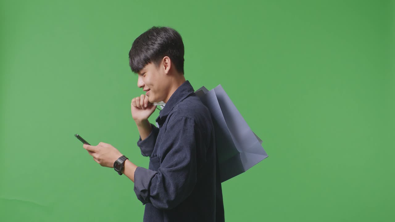 휴대 전화 를 사용 하고 녹색 스크린 앞 에서 는 동안 미소 를 지는 쇼핑 가방 을 들고 있는 아시아인 쇼핑맨 의 측면 시상