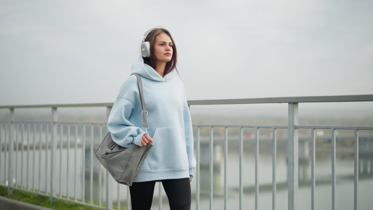 Girl walking confidently with headphones on, holding a bag and a ring in her hand, background features overhead bridge, iron rail, and a river, creating a majestic, urban atmosphere