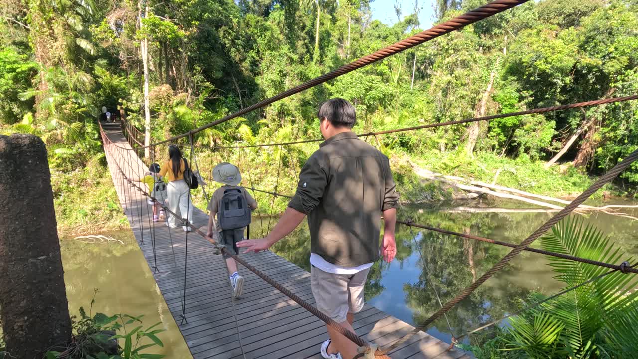 Sebuah keluarga Thailand berjalan melintasi jembatan gantung kayu di atas sungai di hutan hujan lebat.