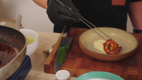 Chef-Masculino-Sin-Rostro-Transfiriendo-Langostinos-Uno-Por-Uno-De-Una-Sartén-Poco-Profunda-Al-Recipiente-De-Chapado-Con-Puré-De-Patata-En-La-Base,-Tiro-Culinario-En-Cámara-Lenta