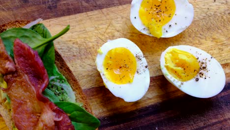 healthy breakfast sandwich on a wooden cutting - avocado, hard boiled eggs, crispy bacon, fresh spinach, salt and pepper, and toasted wheat bread - panning from top down