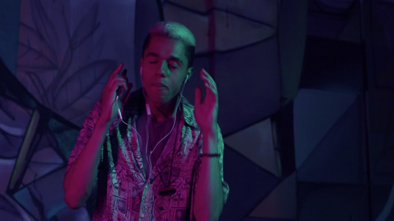 joven afroamericano con estilo escuchando música con auriculares y bailando enérgicamente al aire libre por la noche