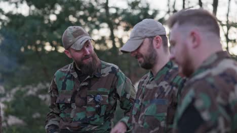 conversación de soldados en ropa de camuflaje, están discutiendo por la noche junto a la fogata común fritando salchichas para la cena, los hombres están descansando después del trabajo, servicio, ejercicios de campo