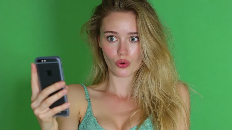 Surprised young woman reacts to something unexpected on her phone, captured against a vibrant green background, showcasing a moment of genuine shock and intrigue