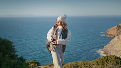 backpack girl taking picture on ocean cliff. happy traveler hiking mountain top