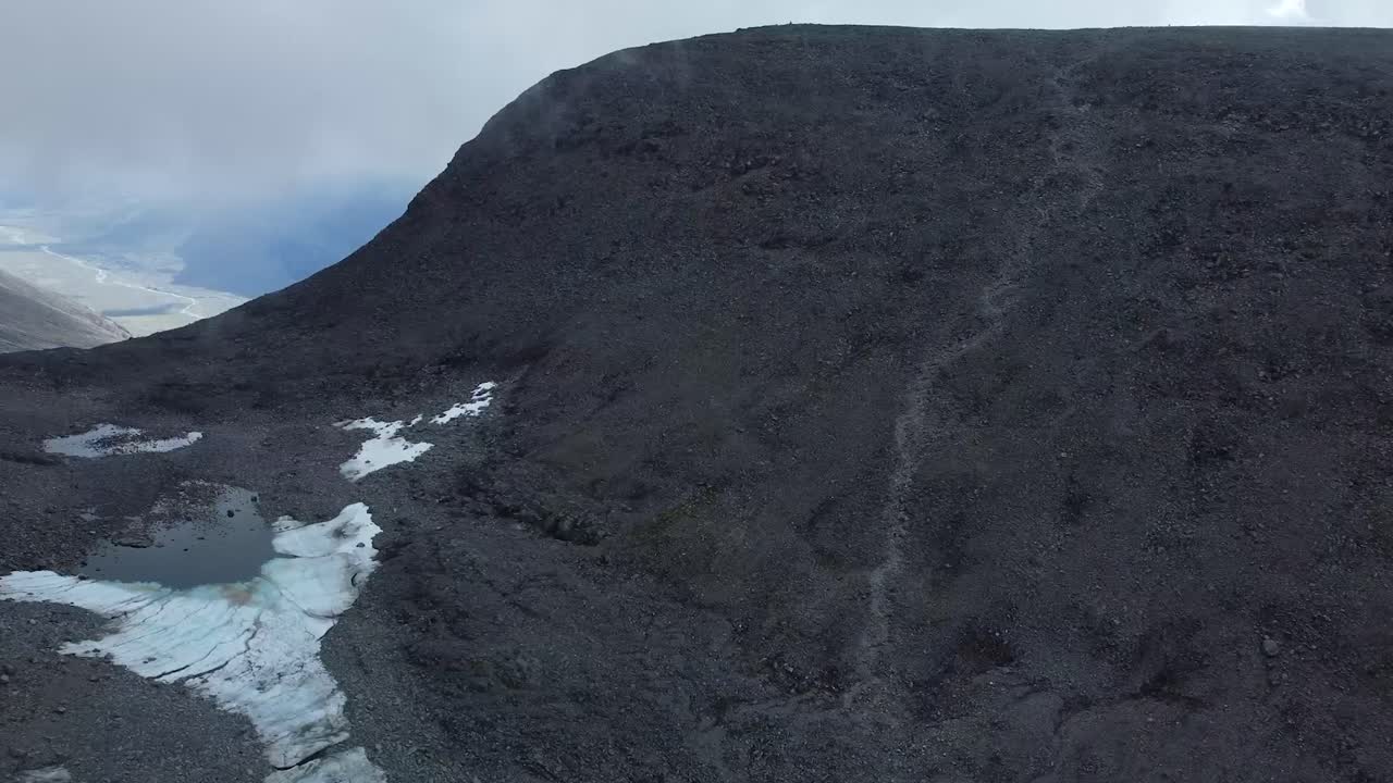 Aerial drone footage view of Large black colored Kebnekaise Volcanic terrain mountain during a cold chilly day with white snow patches on the mountain. Hiking trail leading up the mountain, cloudy day