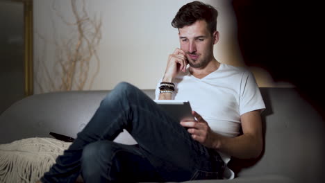 Un-Joven-Con-Barba-De-Varios-Días-Y-Un-Peinado-Moderno,-Vestido-Con-Una-Camiseta-Blanca-Y-Jeans,-Sentado-En-Un-Sofá-En-Casa,-Con-Las-Piernas-Cruzadas,-Riéndose-Mientras-Navega-Por-Su-Tableta,-Estático-4k