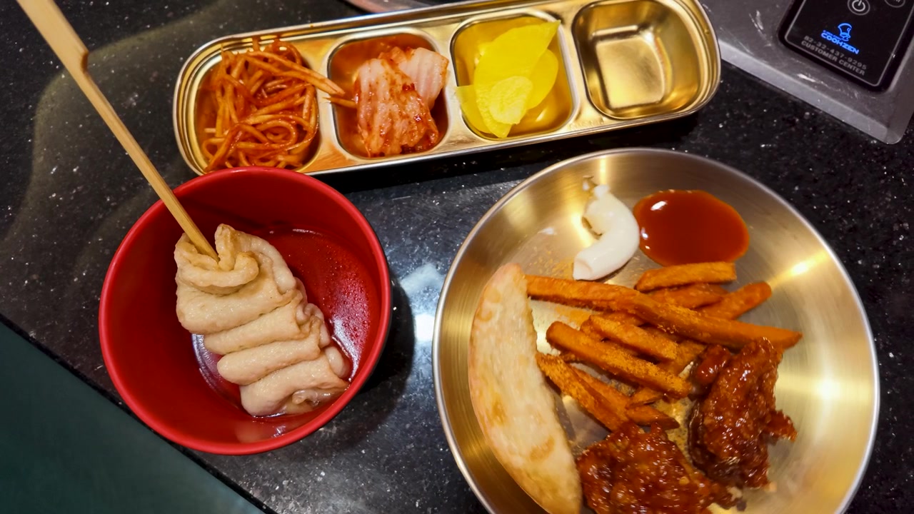 Tampilan atas ayam goreng Korea, kue ikan, dan kimchi yang disajikan di meja restoran