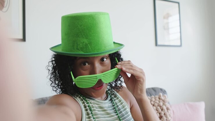 African American woman holding phone, recording selfie at home, adjusting green hat for party