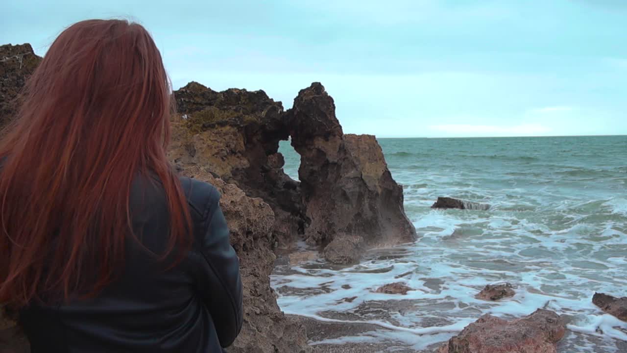 Tranquilla scena diurna di una donna vista da dietro, seduta su una costa rocciosa portoghese mentre osserva potenti onde infrangersi contro le scogliere in un pacifico momento di riflessione
