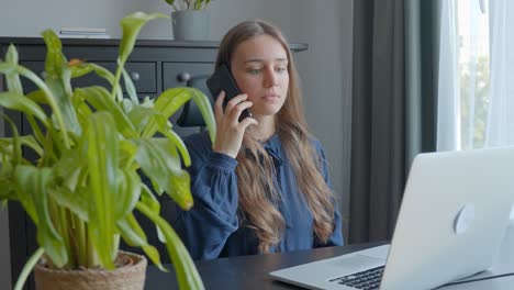 mujer joven sentada junto al escritorio y portátil recogiendo el teléfono móvil