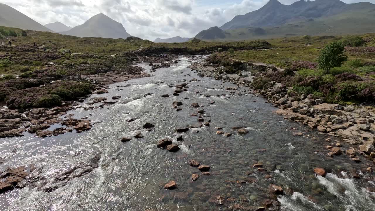 Drone camera smoothly glides above a clear, rocky stream with mountain backdrop on the Isle of Skye, under natural daylight and partly cloudy skies