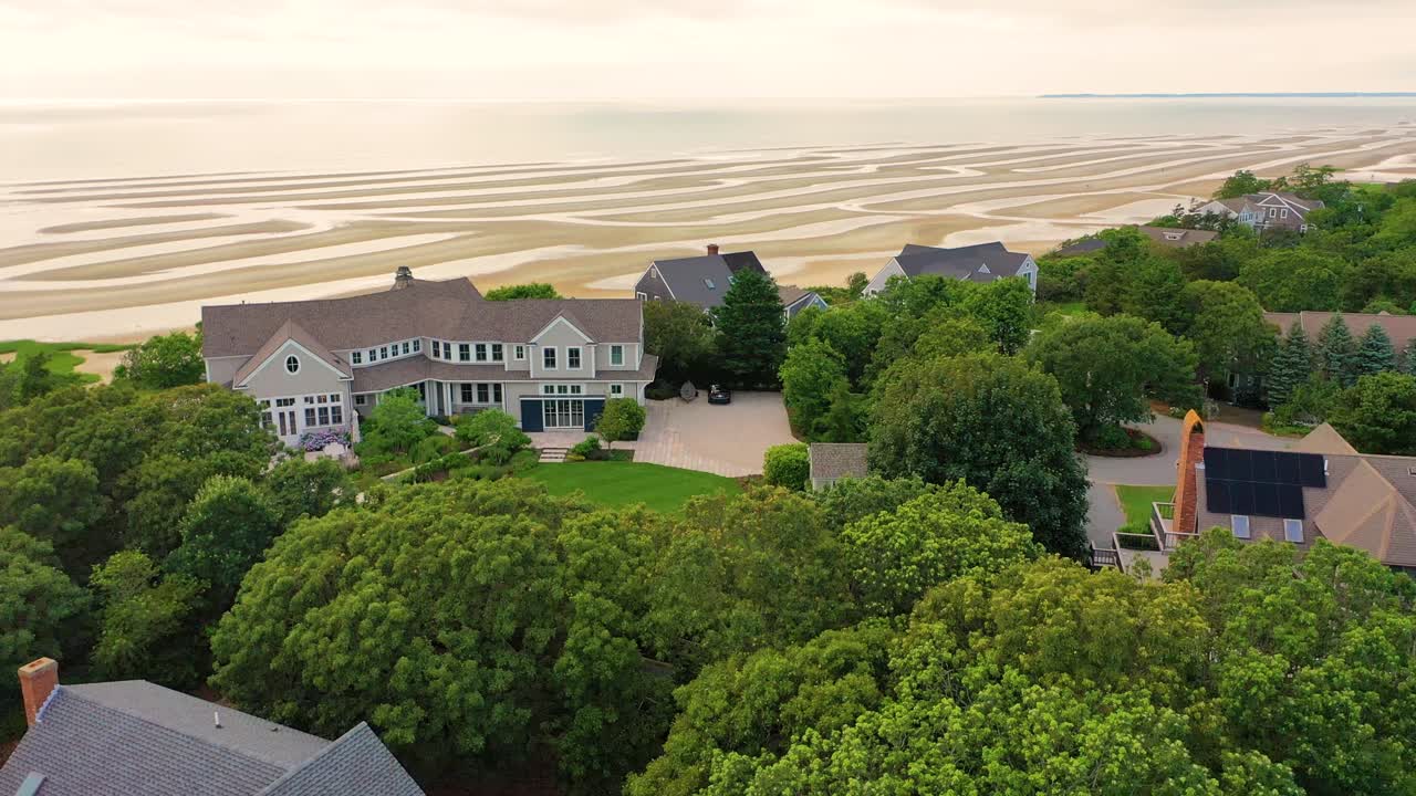 Aerial view of beachfront houses overlooking sandy shoreline and protective stone walls, framed by green dunes and wide marshland stretching across the horizon under a summer sky