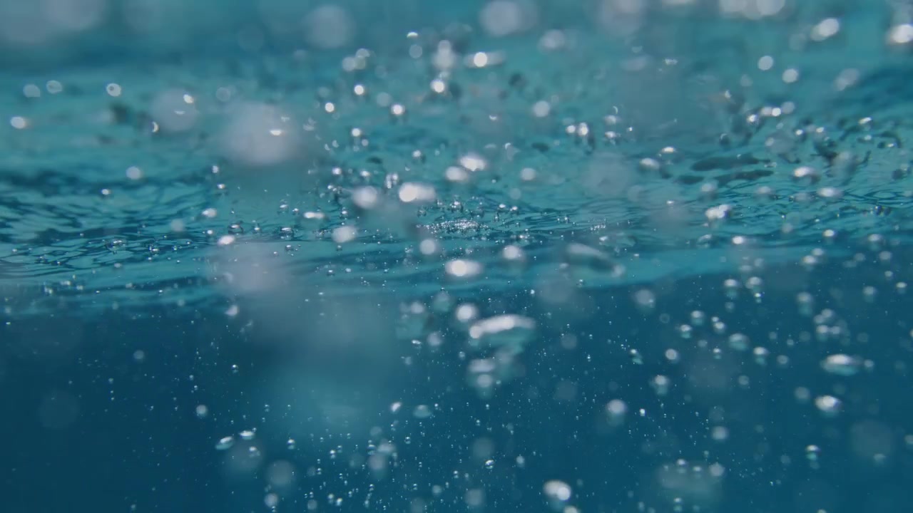 Bolle d'aria salgono sotto la turbolenza della superficie dell'oceano in super slow motion, rivelando cavitazione, taglio dell'acqua e movimento di microbolle sospese attraverso l'acqua di mare blu illuminata.