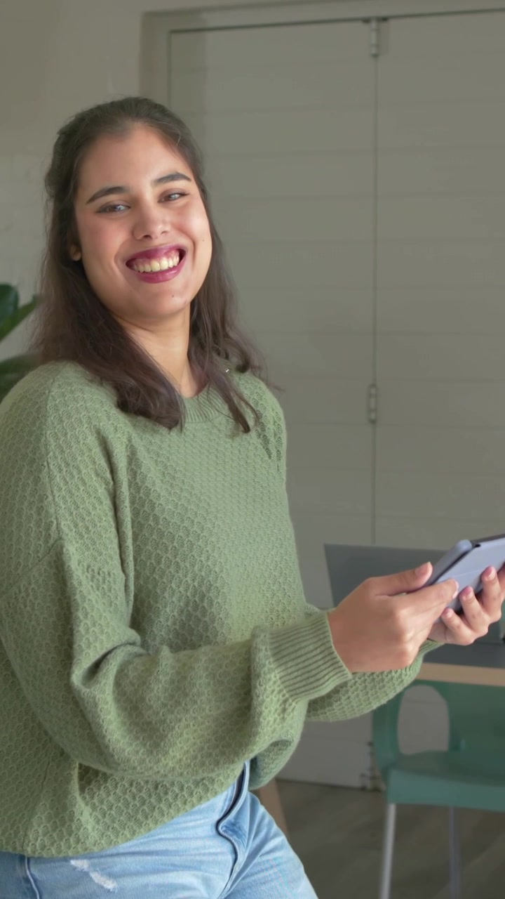 Video verticale: La telecamera si avvicina a una donna con un maglione verde che tiene un tablet a un tavolo in una stanza sorridendo