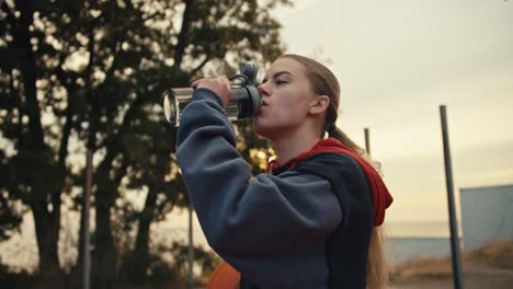 Primer-Plano-De-Una-Chica-Rubia-Con-Una-Sudadera-Con-Capucha-Negra-Bebiendo-Agua-De-Una-Botella-Deportiva-Gris-Mientras-Sostiene-Una-Pelota-De-Baloncesto-En-Su-Mano-Derecha-Durante-Su-Entrenamiento-Matutino-En-La-Cancha-De-Baloncesto-En-El-Verano.