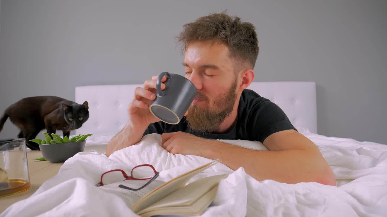 Giovane uomo caucasico a letto sorseggia il caffè mattutino in una camera da letto domestica bianca