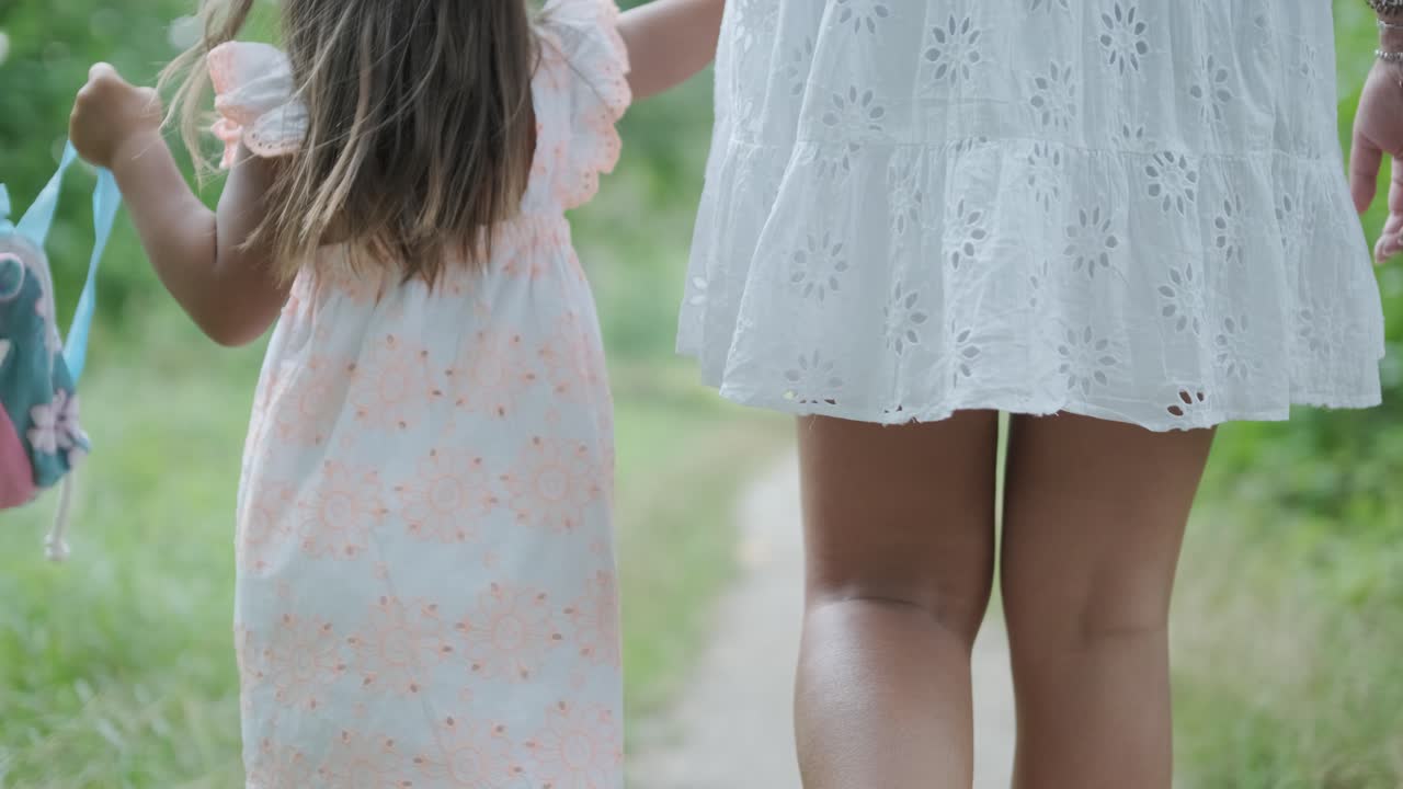 una mujer embarazada y su pequeña hija, ambas vestidas de blanco, caminan de la mano a través de un parque cubierto de hierba. la escena captura un momento sereno de unión familiar en la naturaleza, rodeada de árboles.