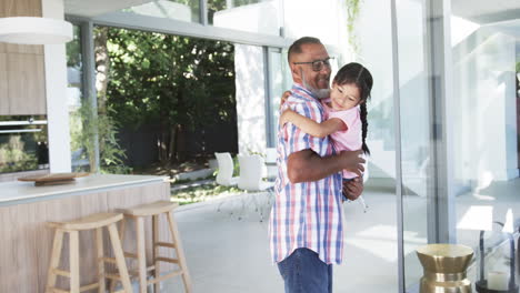 Biracial-grandfather-in-a-plaid-shirt-carries-a-young-biracial-granddaughter-indoors