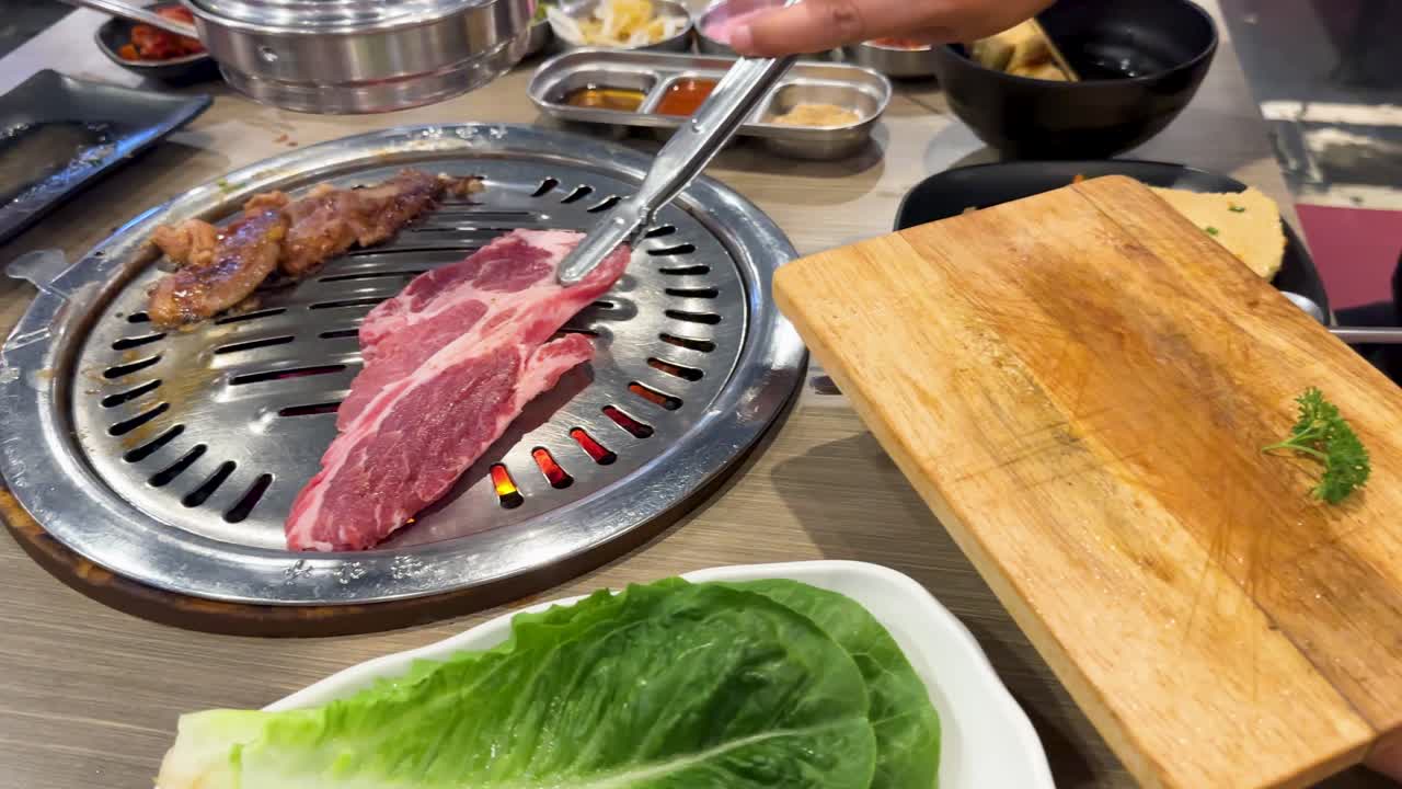 Raw pork placed on a grill in a vibrant Bangkok restaurant. Bright lighting highlights the marbled meat and fresh ingredients