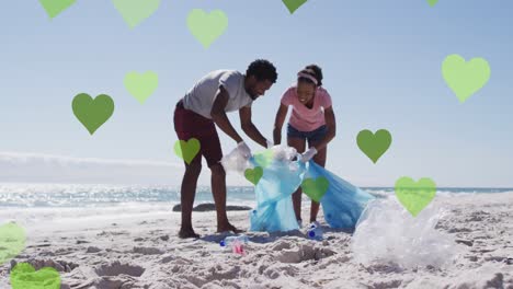 animación de corazones verdes sobre una pareja afroamericana con bolsas de reciclaje limpiando la playa