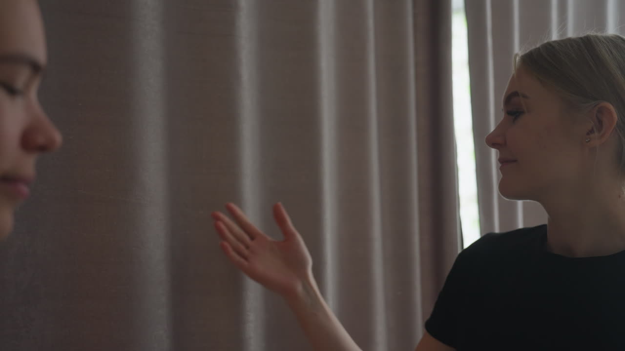 Smiling blonde woman dressed in black gently gestures with hand toward another lady while standing near grey curtain in softly lit interior, creating a moment of warm interaction and quiet expression