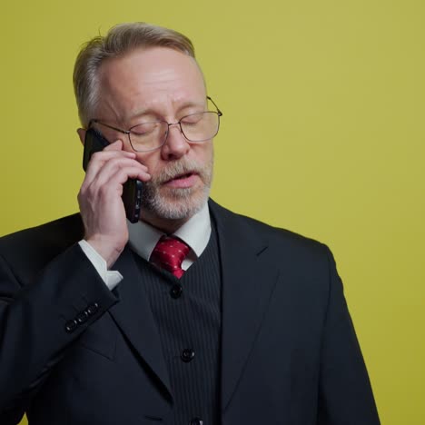 nervous middle aged businessman with a cellphone. mature man in dark suit talking on the phone and giving instructions to somebody on yellow background.