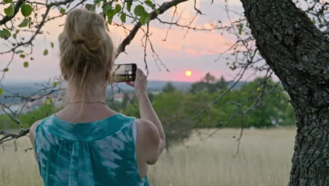 una mujer bajo un árbol toma una foto telefónica del cielo al atardecer de verano en el campo