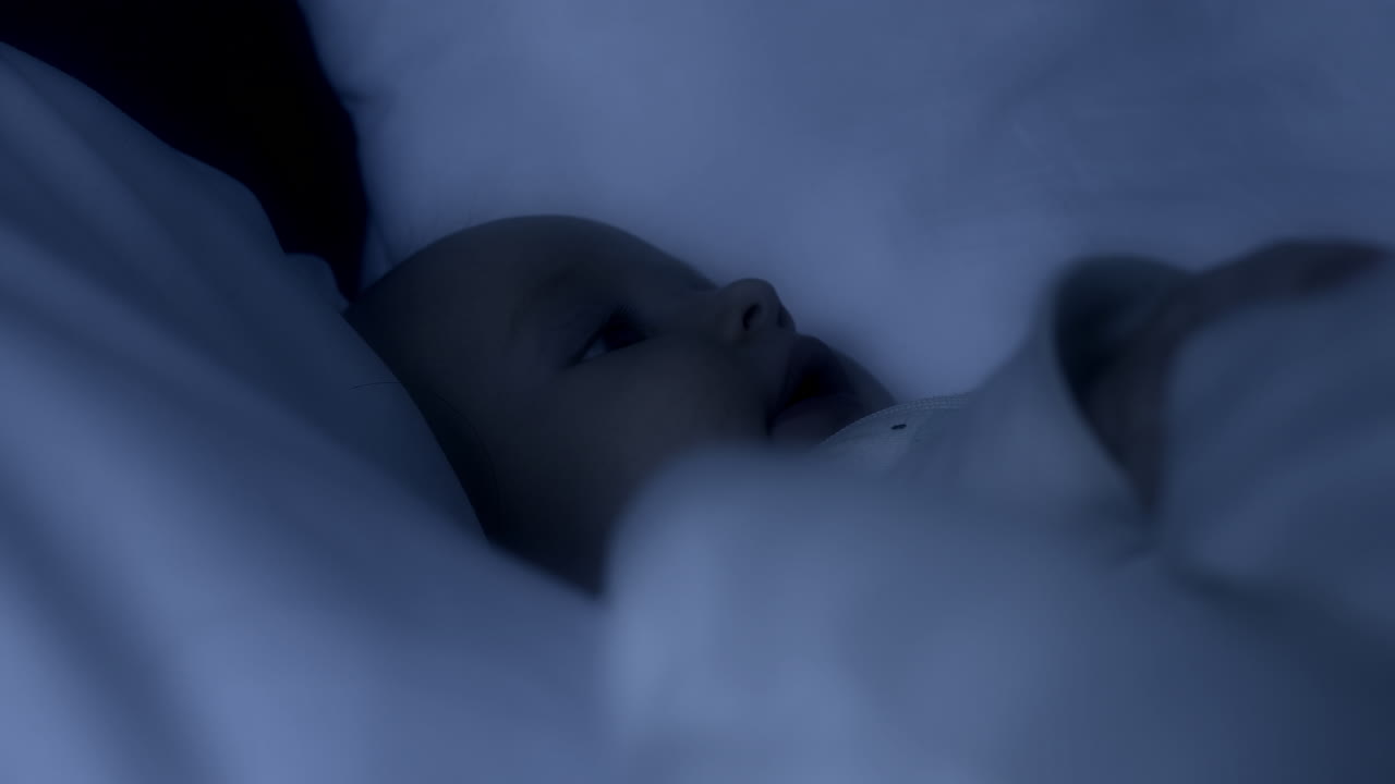 그의 부모의 침대에서 밤에 깨어 있는 아기의 흰색 뭉치의 근접