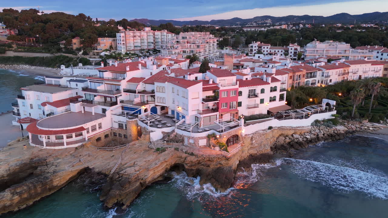 Stunning aerial view of a coastal town in Costa Dorada. The drone ascends, revealing cliffs, turquoise waters, and the charming architecture of this Mediterranean paradise at sunset.