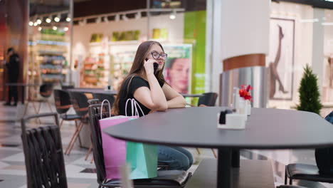 elegant young woman in glasses happily talking on phone, smiling, looking right, seated in modern food court with blurred background featuring structures and tables, with passerby visible in distance