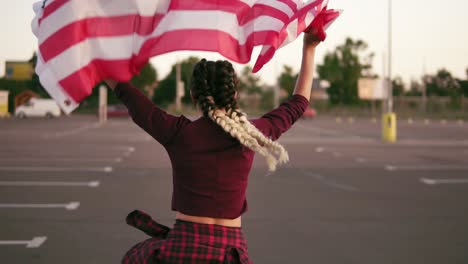 Vista-Posterior-De-Una-Joven-Americana-Feliz-Corriendo-Mientras-Sostiene-La-Bandera-Americana.-Luego-Se-Da-Vuelta-Y-Mira-A-La-Cámara.