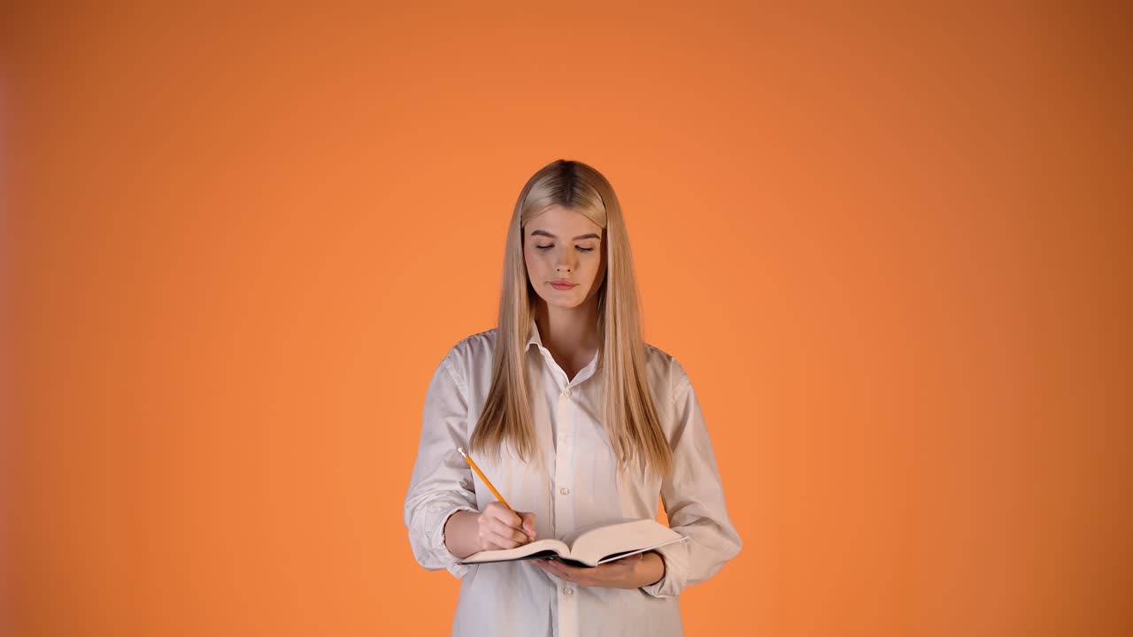Ung blond kvinne skriver notater i notatbok, tenker og finner en smart idé, fargerikt studiobilde