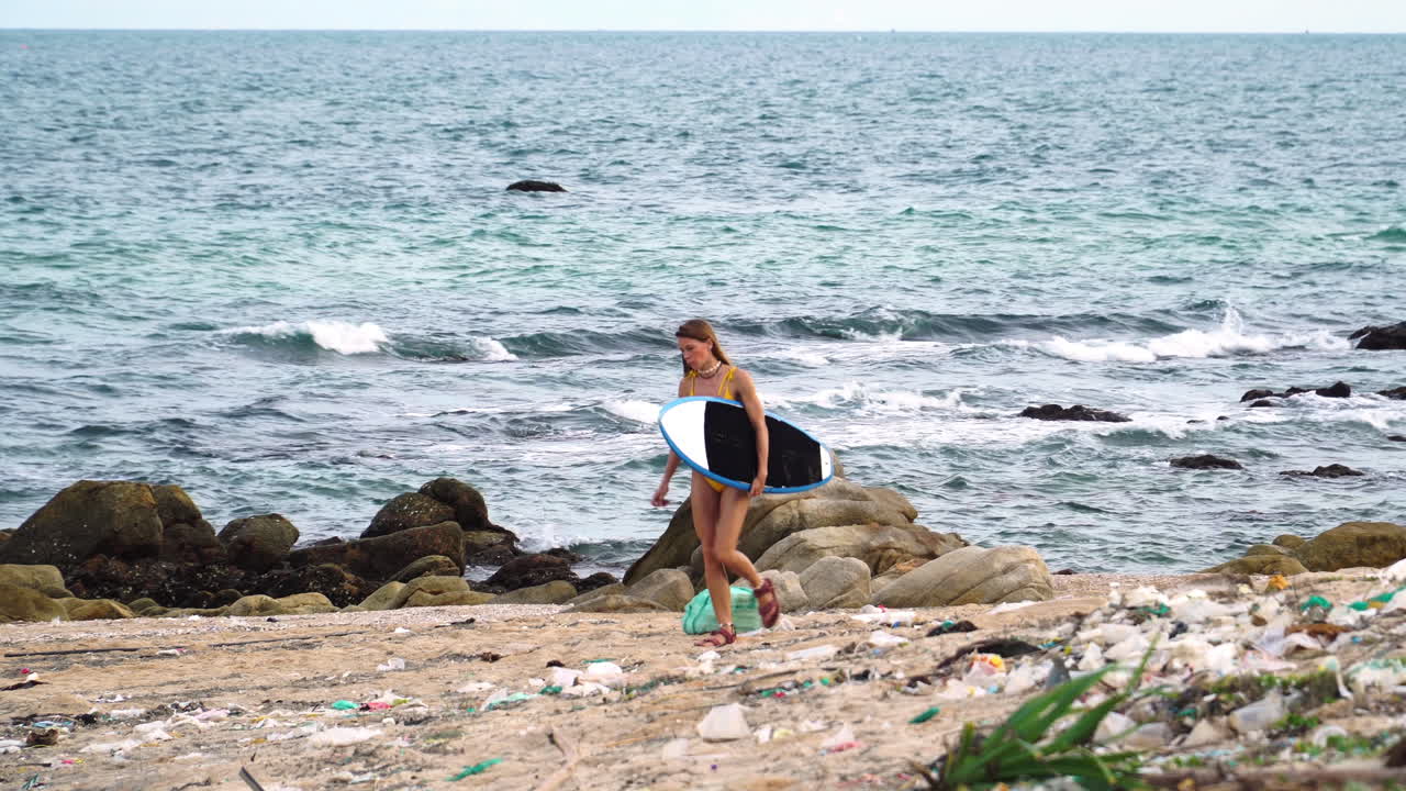 joven surfista en bikini camina en sandalia en la playa con desechos plásticos y contaminación del océano, mujer caminando sostiene una tabla de surf