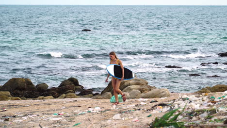 Joven-Surfista-En-Bikini-Camina-En-Sandalia-En-La-Playa-Con-Desechos-Plásticos-Y-Contaminación-Del-Océano,-Mujer-Caminando-Sostiene-Una-Tabla-De-Surf