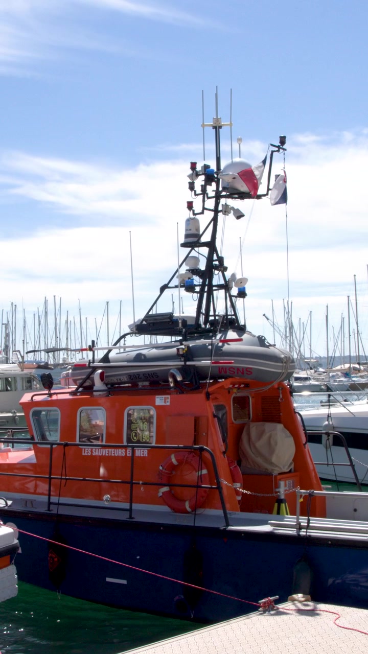 Un'imbarcazione di salvataggio arancione è ormeggiata in un soleggiato porto mediterraneo pieno di yacht di lusso