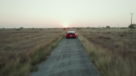 Cinematic-shot-following-red-sporting-car-heading-away-on-the-road-through-the-vast-filed-area-with-low-angle-sunlight