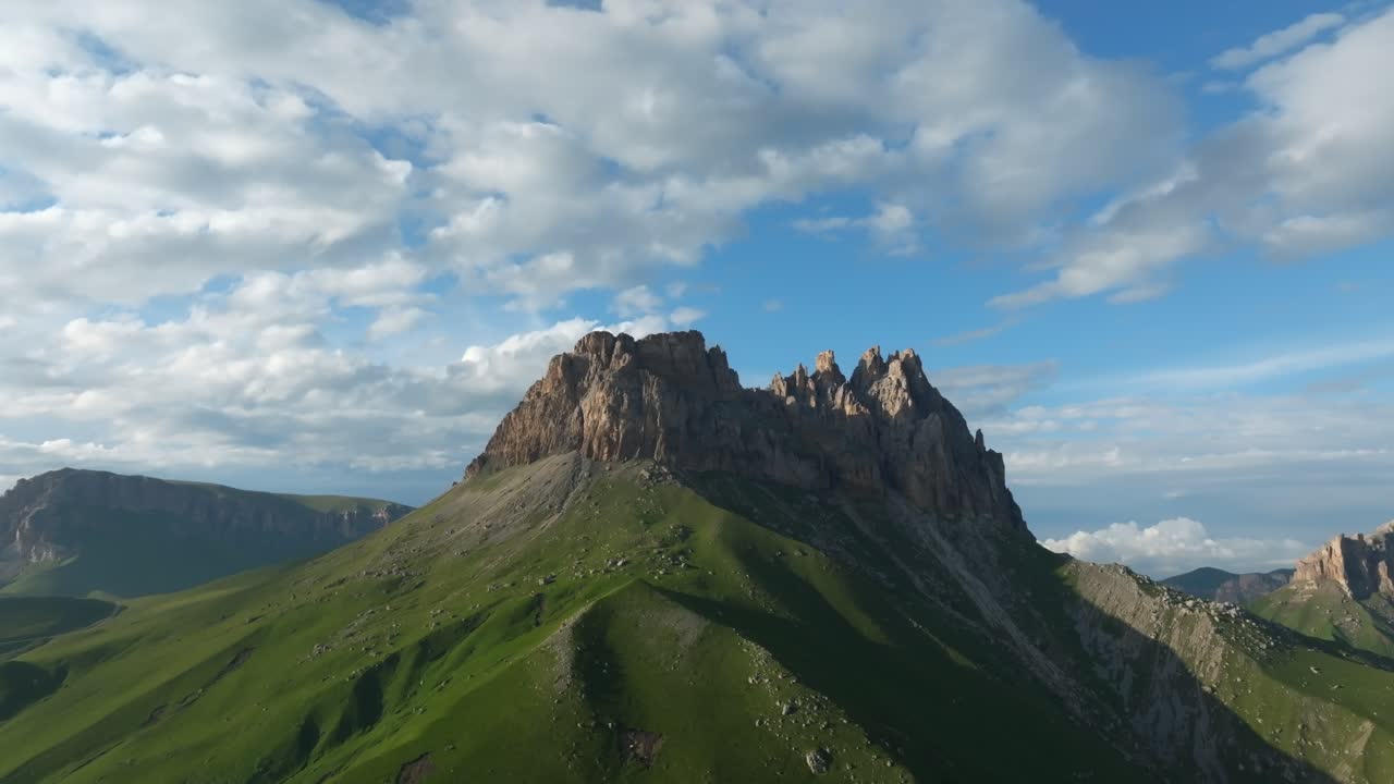 Scenic mountain landscape with jagged rocky summit and lush green slopes under moving clouds, view of Mount Naudzhidza in Russia, wide outdoor nature panorama