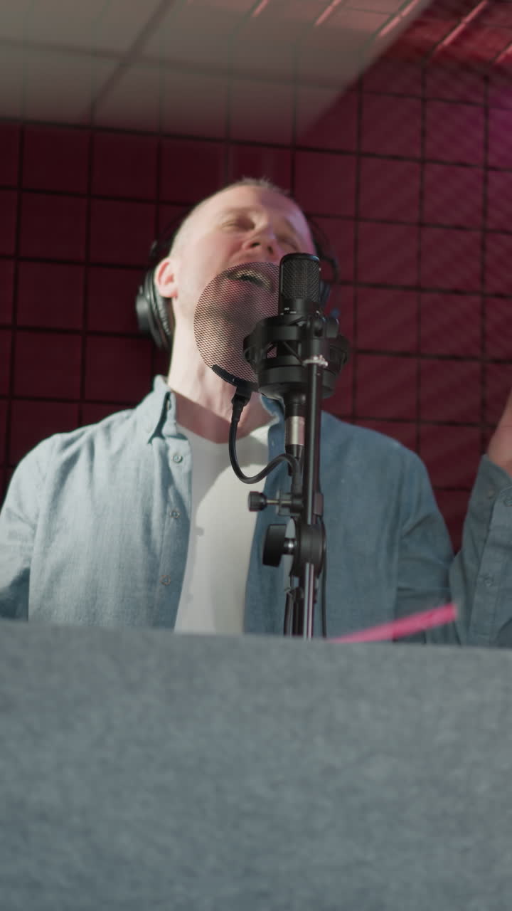 un hombre canta apasionadamente en un estudio de grabación, con auriculares negros y una camisa azul de manga larga. actúa con los ojos cerrados y el movimiento de las manos, rodeado de paneles acústicos rojos