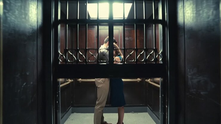 La telecamera si allontana rivelando un uomo che abbraccia una donna in abito blu, condividendo un momento intimo in un ascensore vicino a un cancello.