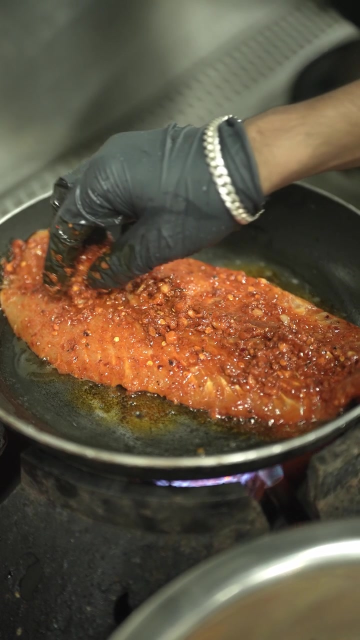 Ripresa verticale ravvicinata di una mano guantata in nitrile nero che spalmA ulteriore marinata piccante rossa su un filetto di pesce già marinato, mentre frigge in olio bollente in una padella di ferro, con un leggero movimento panoramico della telecamera
